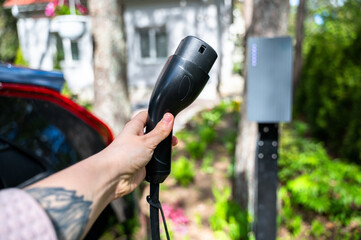 Person grips electric vehicle charger with car partially visible in background, parked in residential yard. Scene reflects shift toward sustainable mobility at home