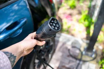 Person grips electric vehicle charger with car partially visible in background, parked in residential yard. Scene reflects shift toward sustainable mobility at home