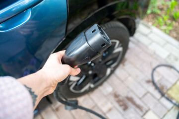 Person grips electric vehicle charger with car partially visible in background, parked in residential yard. Scene reflects shift toward sustainable mobility at home