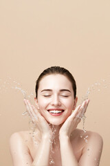 Young woman washing face with water splash on beige background, copy space
