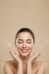 Young woman washing face with water splash on beige background, copy space