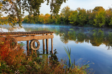 Wooden pier with rubber car tires on a river with trees along the banks. Picturesque landscape of the river in the countryside on a autumn morning