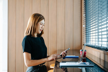 Asian businesswoman on smartphone while reviewing clipboard and laptop in a modern remote office.