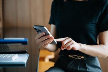 Asian businesswoman using smartphone and laptop in a modern workspace by a window, showcasing digital communication