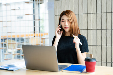 Asian businesswoman on smartphone while reviewing clipboard and laptop in a modern remote office.