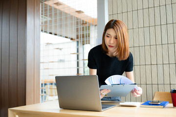 Asian businesswoman calculates finances on a clipboard with calculator by her laptop and smartphone...