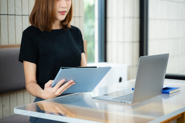 Asian businesswoman calculates finances on a clipboard with calculator by her laptop and smartphone...