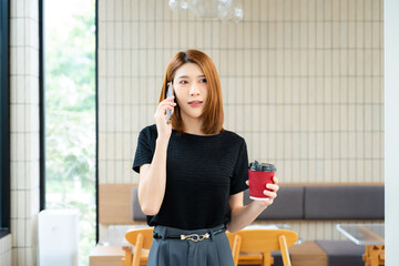 Asian businesswoman talking on smartphone while holding a pen and reviewing a laptop in a modern office