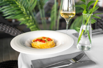 Elegant gourmet ravioli topped with red caviar and herbs, served on a white plate with wine and cutlery in a refined restaurant setting.