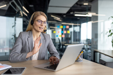 Fototapeta premium A businesswoman in a suit and headset waves during a video call, working from her office in front of a laptop.
