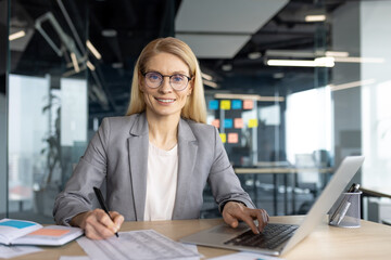 A smiling businesswoman in a modern office setting, working on a laptop while taking notes,...
