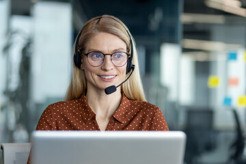 A smiling blond woman wearing a headset and glasses works at a laptop in a modern office setting,...