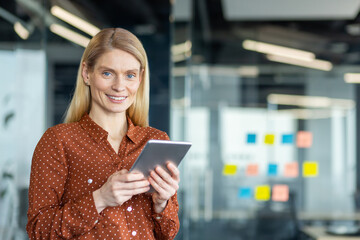 A smiling businesswoman in an office setting holds a tablet, ready for business.