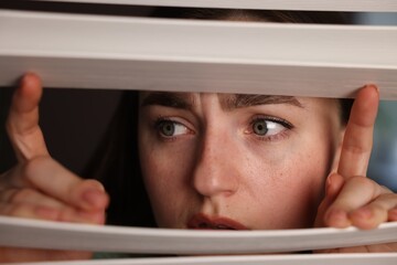 Paranoia. Worried woman looking through window blinds indoors