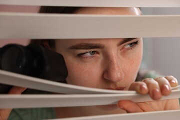 Paranoia. Worried woman with binoculars looking through window blinds indoors