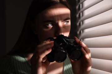 Paranoia. Worried woman with binoculars near window indoors