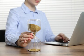 Deadline. Woman with hourglass and laptop working at wooden table in office, closeup