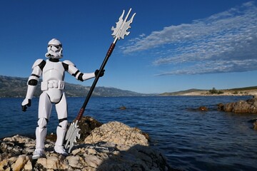 Naklejka premium Star Wars action figure of Clone trooper in white armor and helmet, holding spiked long asian staff in position like Poseidon with triton. Croatian rocks on bay shore, blue skies with clouds.