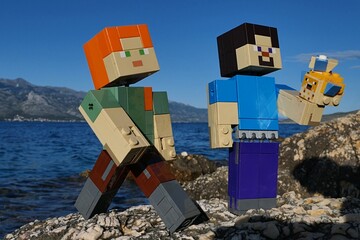 Obraz premium LEGO Minecraft Steve examining LEGO Pufferfishes aquatic mobs, Alex walks next to him. Location rocky Croatian beach, bay water and Velebit mountain in background. Blue summer skies. 