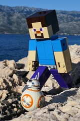 Naklejka premium Closeup view of LEGO Minecraft figure of Steve on walk with small model os Star Wars astromech droid BB-8 on rocky Croatian beach. Bay water, Velebit mountain and blue skies in background. 