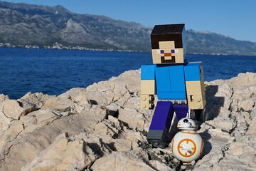 Naklejka premium LEGO Minecraft figure of Steve on walk with small model os Star Wars astromech droid BB-8 on rocky Croatian beach. Bay water, Velebit mountain and blue skies in background. 