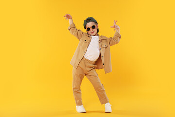 Cute little boy in sunglasses and baseball hat dancing on orange background