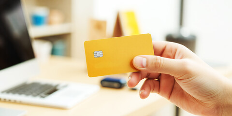 A hand holding a yellow credit card in front of a laptop on a desk in a bright room on transparent background