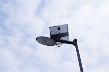Solar panel small with light bulb attached to steel electric pole. On side of road, street sky and...
