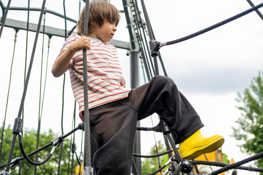 A child boy is climbing up an alpine grid in a park on a playground on a hot summer day. Kids playground in a public park, entertainment and recreation for children, mountaineering training.