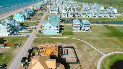 Aerial view residential construction near ocean shoreline with framing work, roofing, heavy equipment, and sandy terrain, vacant land, energy and scale of active development in Galveston, Texas