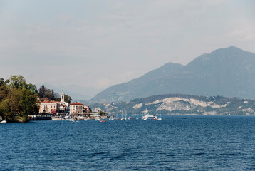 Naklejka premium Scenic Lake Maggiore vista featuring sailboats gliding over azure waters, Belgirate nestled amid dramatic mountain peaks, capturing classic Italian lakeside tranquility