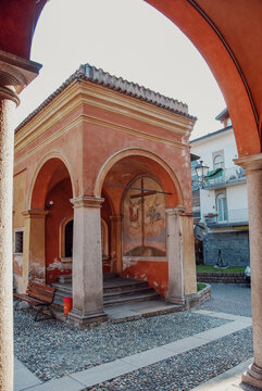 Oratory of San Rocco in Ispra, Lake Maggiore, Italy, showing arches, columns, fresco painting, cobblestone pavement and a glimpse of a modern building on a sunny day