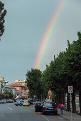 Vivid rainbow arcs over a tranquil street near Lake Maggiore in Piedmont, Italy. Lush trees line...