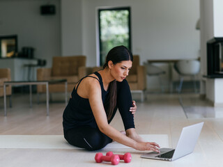 A young woman in sportswear is sitting in the living room and preparing for online training while...