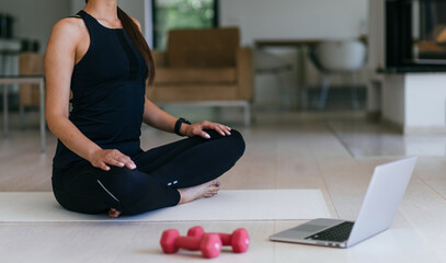 Obraz premium A young woman in sportswear is sitting in the living room and preparing for online training while using a laptop.