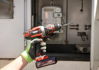 Close-up of a gloved hand holding a cordless electric drill in front of an open electrical panel....