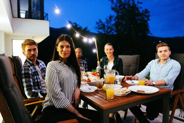 A group of young diverse people having dinner on the terrace of a modern house in the evening. Fun for friends and family. Celebration of holidays, weddings with barbecue