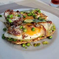 This is a plate of toast topped with sunny-side-up eggs, crispy bacon, and fresh green peas.