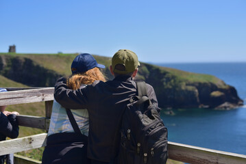 A couple stands on a wooden railing, admiring the coastal scenery of cliffs and the ocean. Husband...