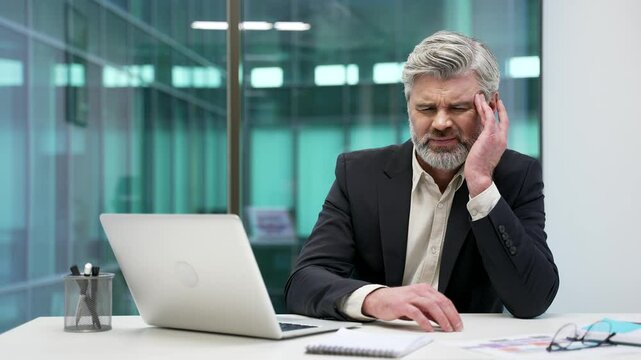 Mature businessman suffering from headache working on laptop while sitting at workplace in office. Unhealthy worker in formal suit holding and massaging his temples with his hands. He is exhausted