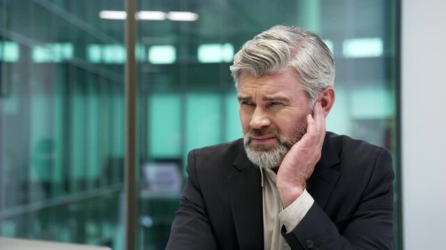 Mature businessman suffers from ear pain sitting in office. Upset male in formal suit touches his ear with fingers. He has furuncle, shingles, eczema, ear diseases or otitis media. Earache. Close up