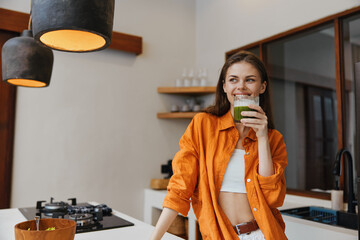 Happy young woman in an orange shirt enjoying a green smoothie in a modern kitchen, emphasizing...