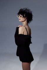 stylish woman wearing sunglasses and black off-shoulder dress posing confidently against gray...