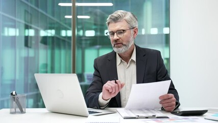 Joyful mature businessman is satisfied with results of a financial report looking at documents and computer screen sitting in business office. Manager in formal suit is happy with positive indicators - Powered by Adobe