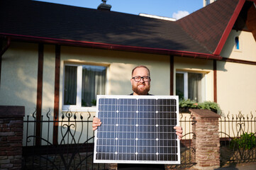 Portrait of happy bearded man with glasses stands in front of house, proudly holding solar panel,...