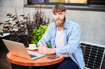 Portrait of bearded man works at round wooden table with laptop, notebook, and cup of coffee. Guy...