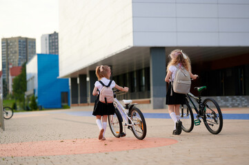 schoolgirls with bicycles walking to school in urban setting, wearing uniforms and backpacks. early...