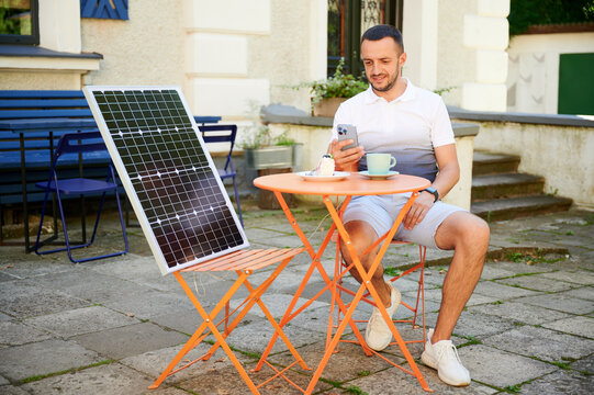Young man charging smartphone from solar panel while sitting in cafe. Handsome male drinking coffee and checking his phone next to solar battery. Conscious male choosing sustainable lifestyle. - Powered by Adobe