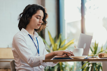 Young woman typing on laptop during lunch break in bright office cafeteria with coffee