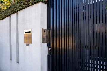 Modern residential entrance of a contemporary home featuring a sleek black slatted security gate, intercom, high garden boundary concrete wall, and mailbox.